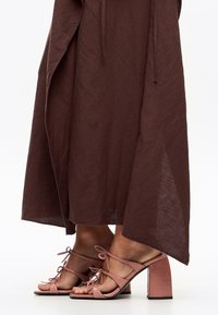 Des pieds chaussés de sandales à talons hauts roses �à lanières, assorties à une longue jupe marron fluide, le tout sur un fond blanc uni.