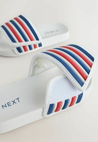 Sandale albe tip papuc cu o bandă lată cu dungi roșii, albe și albastre, talpă texturată și marcate cu "NEXT" albastru.