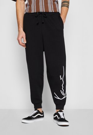 Mann trägt schwarze Jogginghose mit weißem Signatur-Logo am linken Bein, schwarz-weiße Sneakers und braun gestreiftes Hemd.