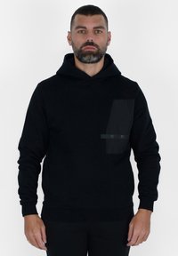 Felpa nera con cappuccio realizzata in tessuto morbido, caratterizzata da una grande tasca frontale con texture liscia e un dettaglio a strisce orizzontali. Polsini e orlo a coste.