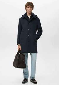 Abrigo de lana azul marino con cuello con cremallera, botones delanteros y dos bolsillos laterales. Combinado con denim claro y un bolso de ante oscuro.