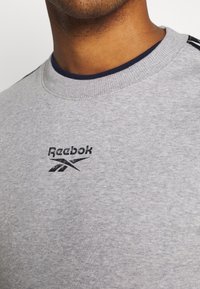 Sweat-shirt gris en tissu texturé. Présente un logo Reebok noir et un col bleu marine. Le design comprend des rayures horizontales subtiles sur les épaules.