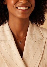 Gouden ketting met een ovale rode edelsteen omringd door kleine heldere stenen, gedragen over een beige gestreepte blazer.