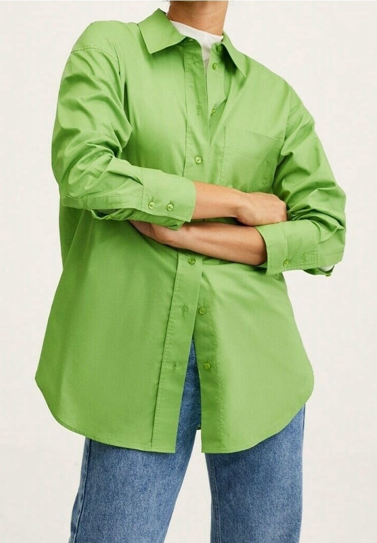 Personne les bras croisés, portant une chemise boutonnée vert vif et un jean bleu sur un fond clair uni.