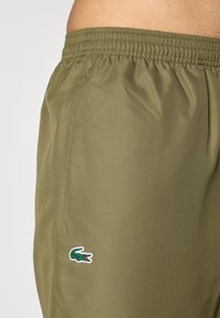 Olivenfargede shorts laget av lett materiale, med elastisk midjebånd og et hvitt og grønt Lacoste-logo-patch på venstre side.