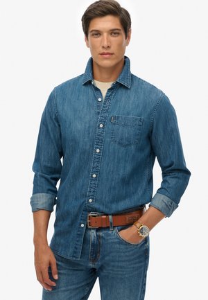 Camisa de mezclilla en azul medio, diseño de botones con un bolsillo en el pecho izquierdo. Combinada con jeans azules y un cinturón marrón. Reloj de acero inoxidable visible.