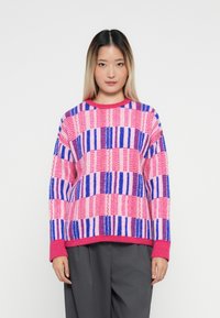 Young woman with straight ombre hair wearing a pink, purple, and blue checkered sweater and dark gray pants, standing against a white background.