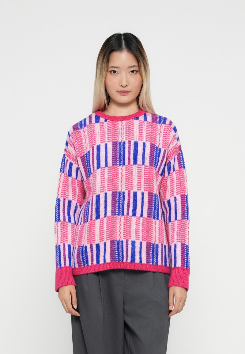 Young woman with straight ombre hair wearing a pink, purple, and blue checkered sweater and dark gray pants, standing against a white background.