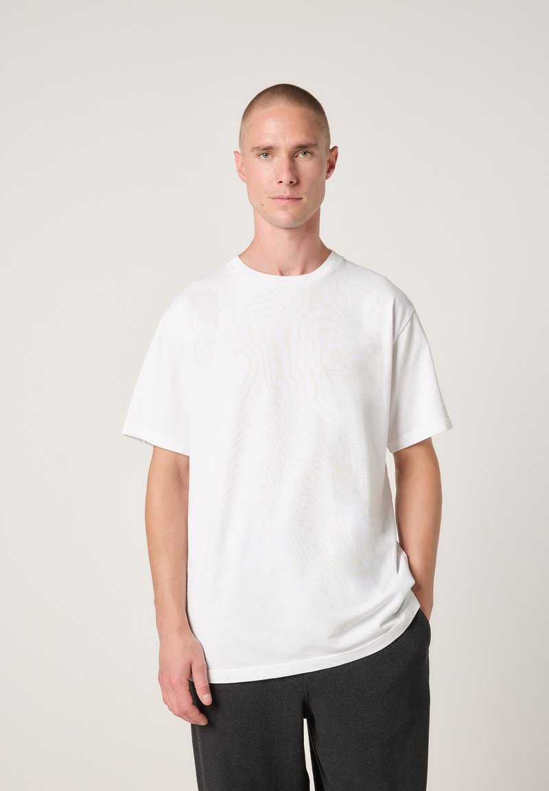 Weißes Baumwoll-T-Shirt mit kurzen Ärmeln, rundem Ausschnitt und lockerem Schnitt. Getragen mit dunkelgrauen Hosen vor neutralem Hintergrund.