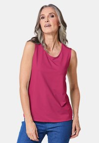 Fuchsia mouwloze top met een ronde hals, gemaakt van gladde stof. Gecombineerd met blauwe jeans poseert het model met één hand op haar heup.