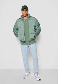 Chaqueta acolchada verde con cuello alto, combinada con una camiseta verde a juego, jeans de mezclilla azul claro y zapatillas blancas. Diseño simple y textura suave.