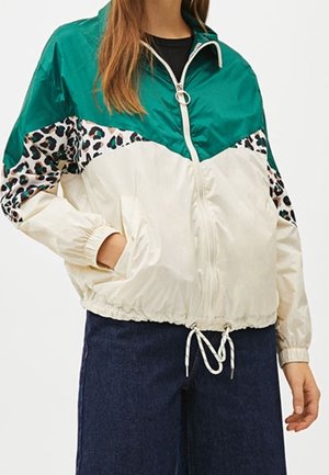 Vrouw die een color-block windjack draagt met groene, witte en luipaardprintpanelen, gecombineerd met donkerblauwe wijde broek.