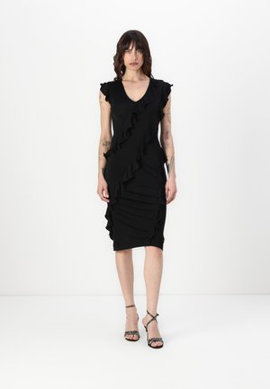 Black dress with ruffle details along the front and shoulders, fitted shape, knee-length, made from smooth fabric. Black strappy heels.