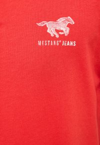 Red cotton t-shirt featuring a white graphic of a running horse and the text "MUSTANG® JEANS" in a bold font below.