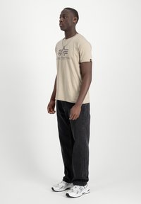 Camiseta de manga corta beige con un estampado de logo en el pecho, combinada con jeans negros y zapatillas deportivas blancas. Diseño sencillo, corte regular.