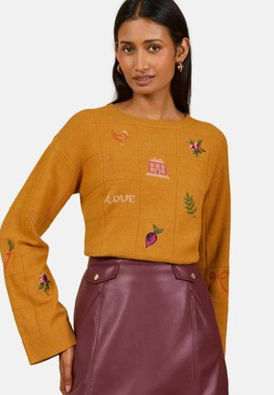 Femme portant un pull jaune moutarde avec des motifs et des lettres brodés, associé à une jupe en cuir bordeaux taille haute.
