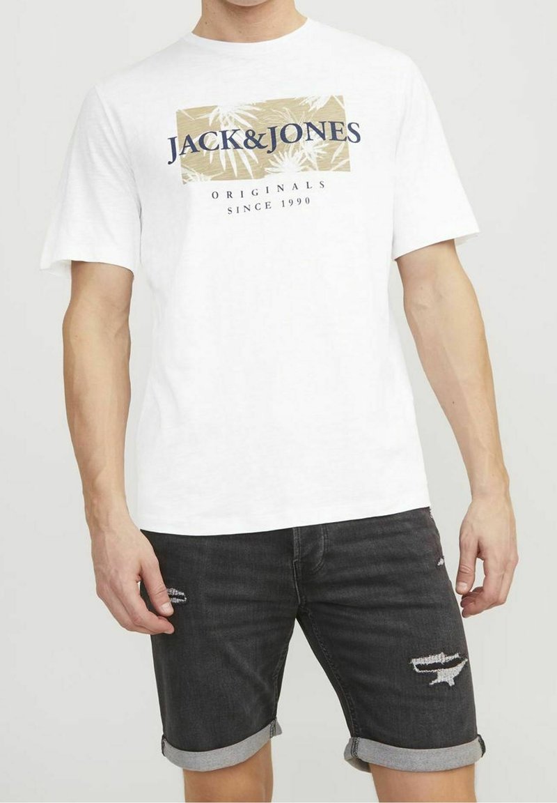T-shirt en coton blanc avec un logo rectangulaire beige et marine, comportant des feuilles de palmier. Porté avec un short en denim noir légèrement déchiré.