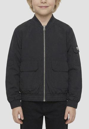 Dijete nosi crnu bomber jaknu na patent s elastičnim manžetama i rubom, dva prednja džepa s klapnom te mali džep na lijevom rukavu.