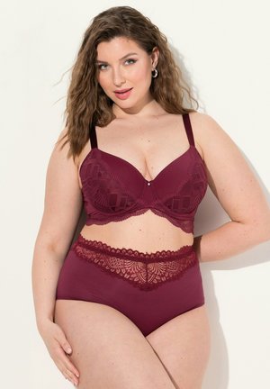 Vrouw met golvend bruin haar die een bijpassende bordeauxrode kanten bh en hoge slip draagt, staand tegen een lichte achtergrond.