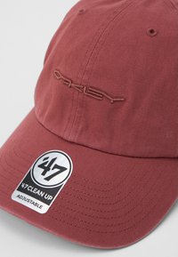 Rdeča bombažna kapa s ukrivljenim obodom, na kateri je izvezen logotip "Oakley" in črna ter srebrna prilagodljiva oznaka spredaj.
