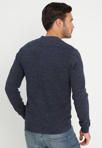 Marinblå stickad tröja med en texturerad mönster, rund halsringning och ribbade ärmslut och nederkant, parat med ljusblå jeans.