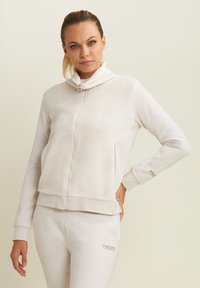 Giacca leggera beige con zip e colletto alto, dalla texture morbida e due tasche laterali. Nome del marchio "FREDDY" ricamato sulla manica.