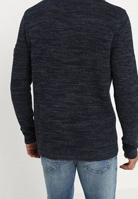 Mörkblå stickad tröja med en strukturerad mönster, rundad halsringning och långa ärmar, kombinerad med ljusblå jeans. Bakvy visas.