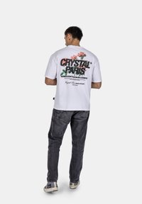 Bílá bavlněná tričko s grafickým designem na zadní straně, které obsahuje "CRYSTAL PARIS" a květinové prvky v černé a růžové barvě. Nosené s šedými džíny.