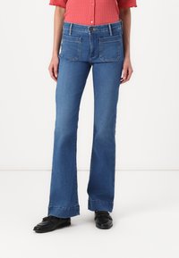 Jean bleu évasé avec une taille haute, en denim, présentant deux poches arrière et un ourlet légèrement effiloché, associé à des chaussures noires.