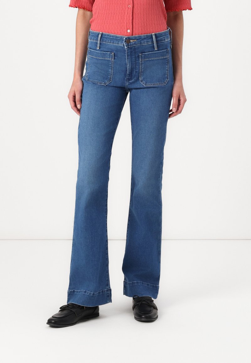 Jean bleu évasé avec une taille haute, en denim, présentant deux poches arrière et un ourlet légèrement effiloché, associé à des chaussures noires.