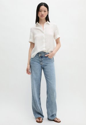 Kobieta stojąca, ubrana w białą koszulę na krótki rękaw z guzikami, jasnoniebieskie szerokie dżinsy i brązowe sandały na gładkim jasnym tle.