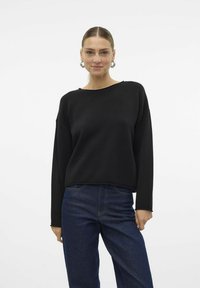 Pull noir surdimensionné en tissu doux avec un décolleté bateau, des manches longues et une coupe décontractée. Associé à un pantalon en denim bleu foncé.