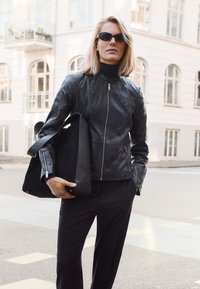 Vrouw die een zwarte leren jas, zwarte coltrui, zonnebril en zwarte broek draagt, met een grote zwarte tas in haar hand, staand op een stadsstraat.