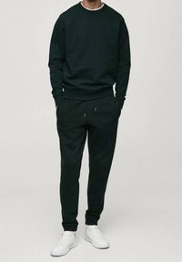 Mężczyzna ubrany w ciemnozieloną bluzę i dopasowane spodnie dresowe, w białych sneakersach, stoi z rękami w kieszeniach na jednolitym tle.