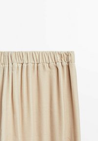 Beige sammet kjol med drar i elastisk midjeband, hängande mot en plain vit bakgrund.