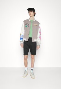 Veste universitaire grise et blanche avec des écussons, t-shirt vert, shorts noirs et baskets montantes blanches. Design minimaliste, style décontracté.