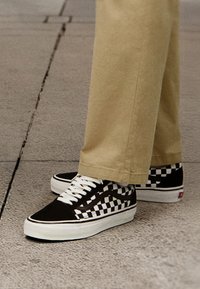 Zapatillas de lona en blanco y negro con un diseño de tablero de ajedrez, con cordones blancos y suela de goma, combinadas con pantalones beige, sobre una superficie gris texturizada.