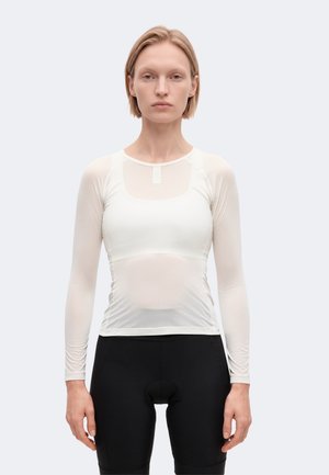 Une femme porte un maillot de cyclisme blanc à manches longues et un short de cyclisme noir sur un fond uni et clair.