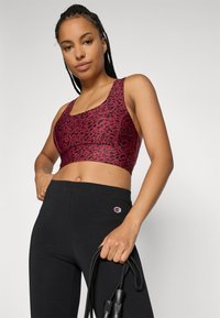 Kvinne i rød leopardmønstret sports-bh og svarte Champion tights som holder et svart hoppetau mot en ensfarget bakgrunn.