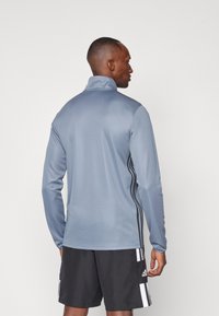 Camisa atlética de manga larga gris con cuello alto, con franjas laterales negras y una textura suave. Usada con pantalones cortos negros.