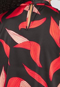 Gros plan sur un tissu noir avec de grands motifs de feuilles rouges et roses, et une petite ouverture en forme de trou de serrure avec un bouton au niveau de l'encolure arrière.