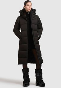 Sort lang puffer-frakke med quiltet design, hætte og lynlås, parret med sorte isolerede støvler med snørebånd fortil og blanke detaljer.