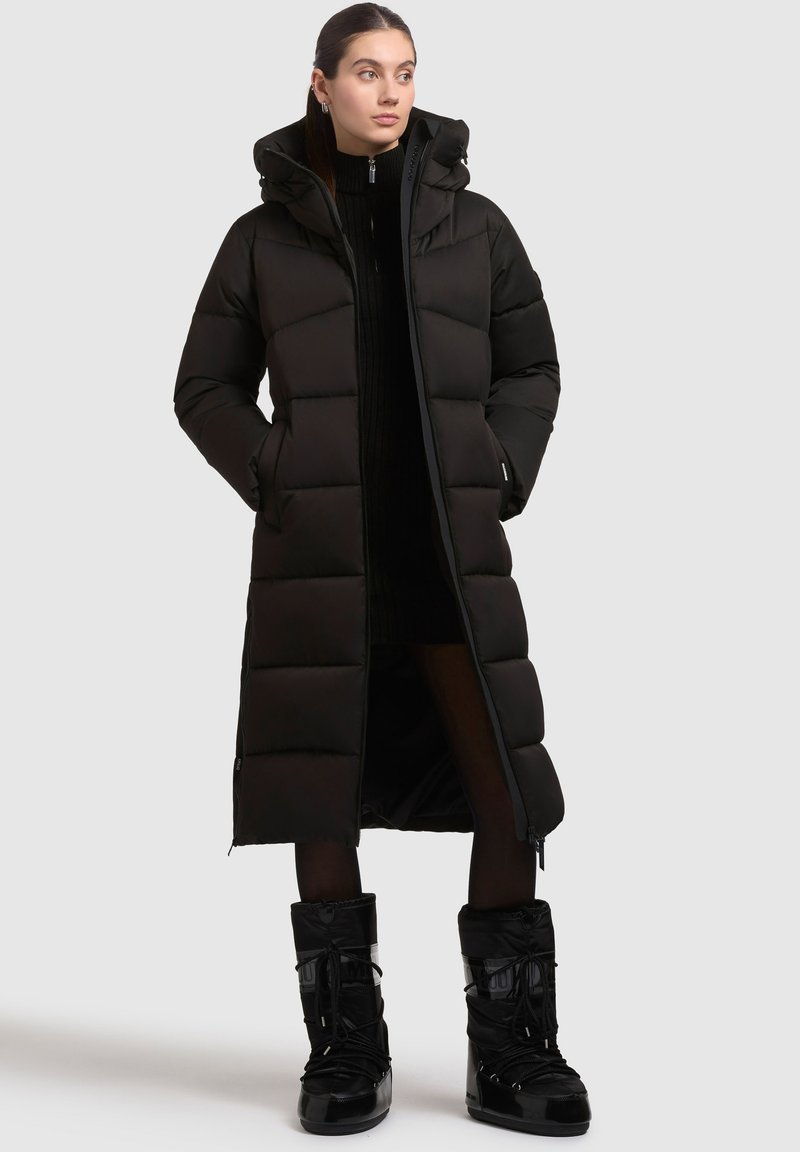 Sort lang puffer-frakke med quiltet design, hætte og lynlås, parret med sorte isolerede støvler med snørebånd fortil og blanke detaljer.