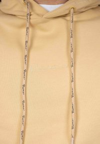 Felpa beige con superficie texturizzata, cordini con testo del logo nero e branding ricamato discreto sul davanti.