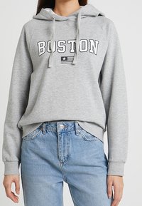Szara bluza z kapturem, z napisem "BOSTON" w grubych czarnych literach, małą grafiką flagi oraz ściągaczami na rękawach i dole. Połączona z jasnoniebieskimi dżinsami.
