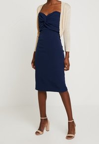 Námořnická midi šaty s dekoltem ve tvaru srdce a zkrouceným detailem na prsou, doplněné lehkým béžovým kardiganem a šlapkami na vysokém podpatku v barvě těla.