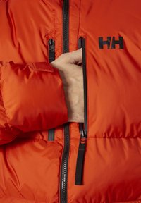 Oranžová zateplená bunda s černým zipem a logem. Obsahuje ruku sahající do zipové kapsy, která ukazuje hladkou texturu a elegantní design.