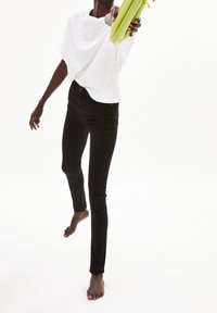 Jeans ajustados negros y una camiseta blanca holgada. El modelo está descalzo, sosteniendo apio con un brazo extendido. Fondo brillante y minimalista.