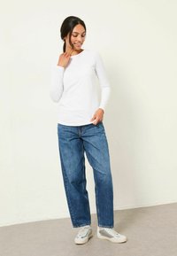Weißes Langarmoberteil kombiniert mit lockeren blauen Jeans und grauen Sneakers, vor einem neutralen Hintergrund.