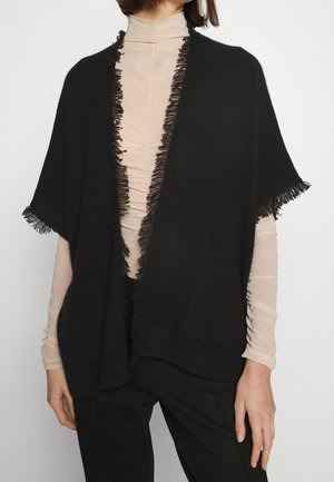 Personne portant un haut à col roulé beige transparent sous un cardigan noir à manches courtes avec des franges sur les bords et un pantalon noir, debout contre un fond blanc.